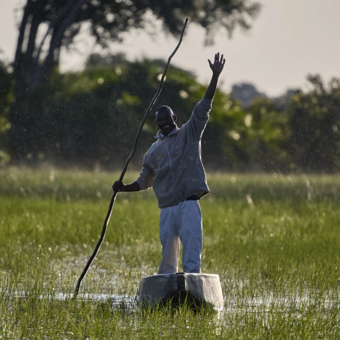 Lächelnder Mann steht mit einer langen Stake in einem flachen Boot im überschwemmten Gras und winkt, Botswana
